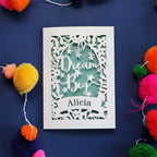 A personalised laser cut congratulations card with the words 'Dream Big' and a name 'Alicia' written on it, displayed on a dark background surrounded by colourful pompons.