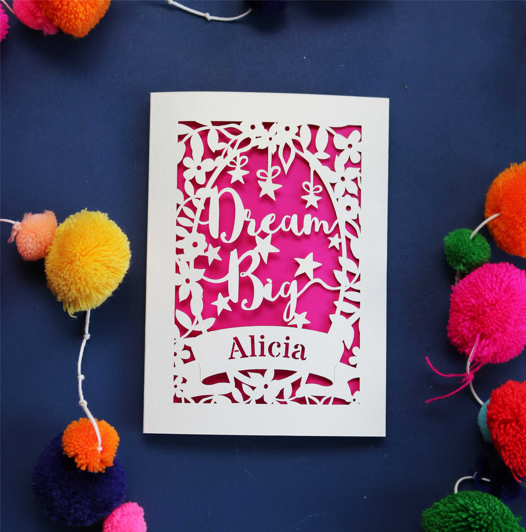 A personalised laser cut card with the words 'Dream Big' and a name 'Alicia' written on it, displayed on a dark background surrounded by colourful pompons.