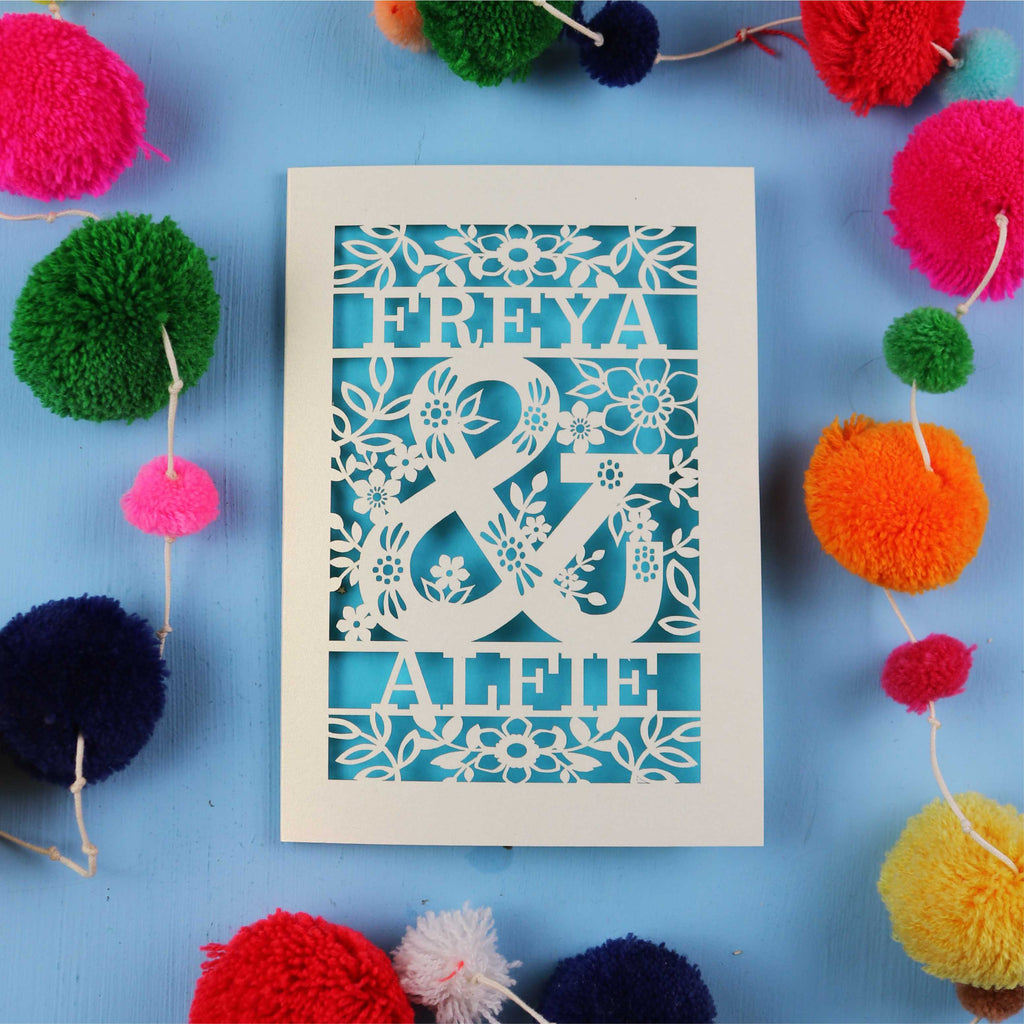Decorative paper cut out engagement card with names 'FREYA' and 'ALFIE' surrounded by colourful pom-poms on a blue background
