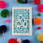 Decorative paper cut out engagement card with names 'FREYA' and 'ALFIE' surrounded by colourful pom-poms on a blue background