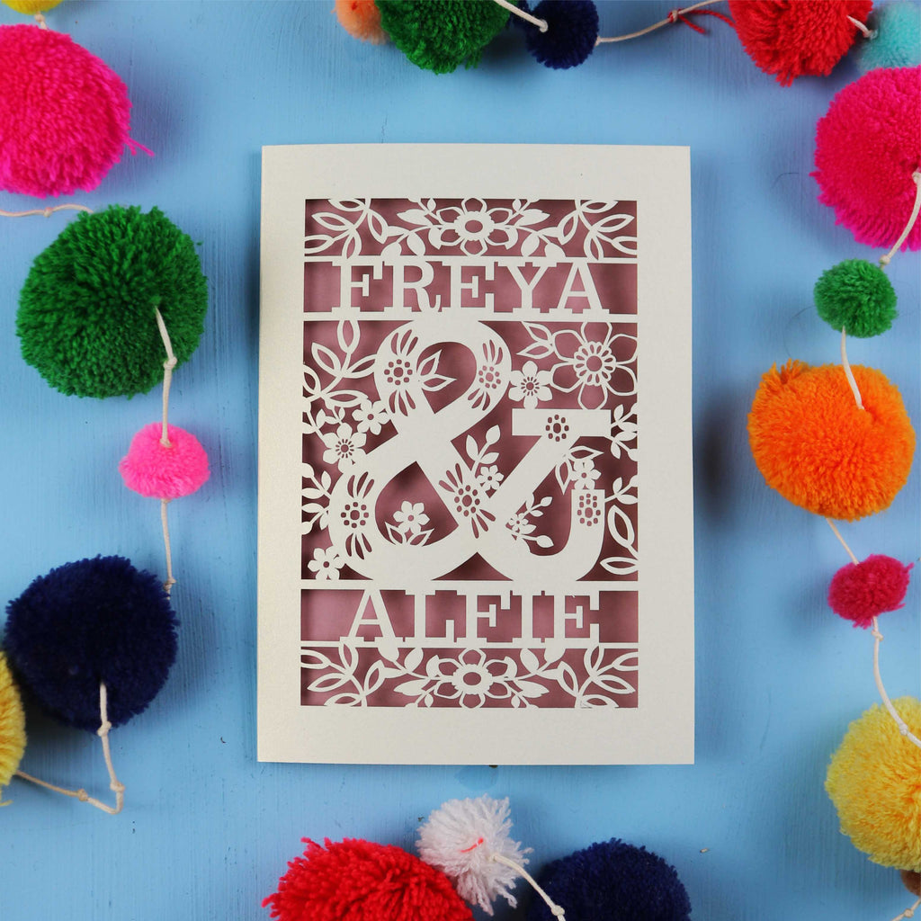 Decorative paper cut out engagement card with names 'FREYA' and 'ALFIE' surrounded by colourful pom-poms on a blue background