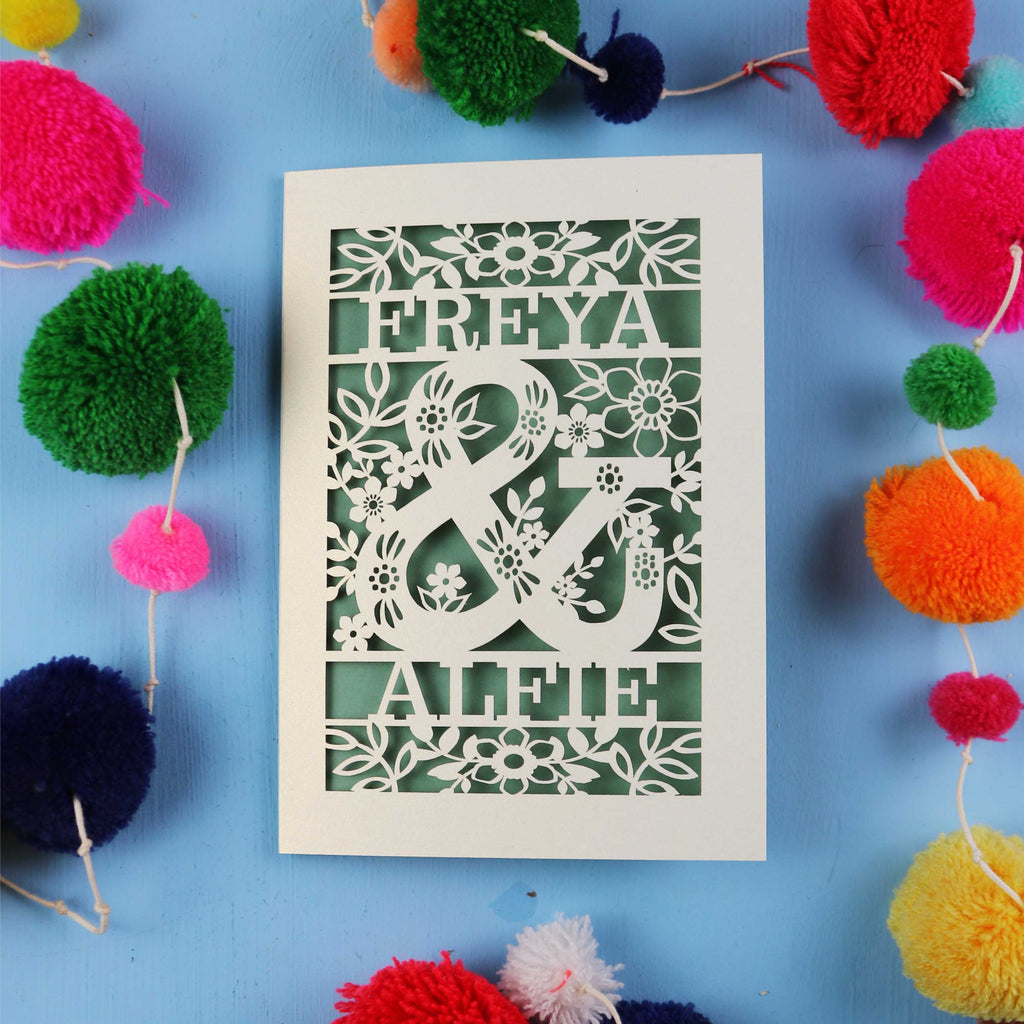 Decorative paper cut out engagement card with names 'FREYA' and 'ALFIE' surrounded by colourful pom-poms on a blue background