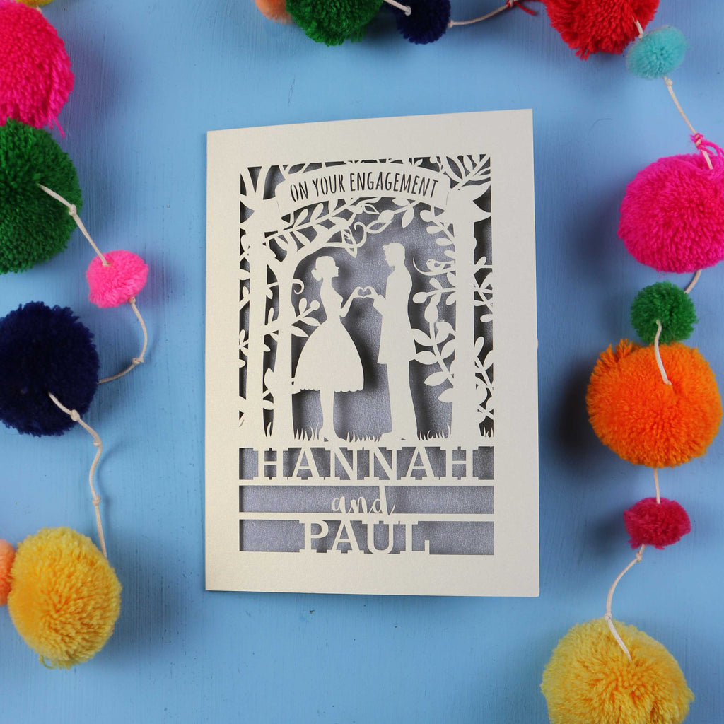 Engagement card with cut-out silhouettes of a couple on a blue background with colourful pom-poms.