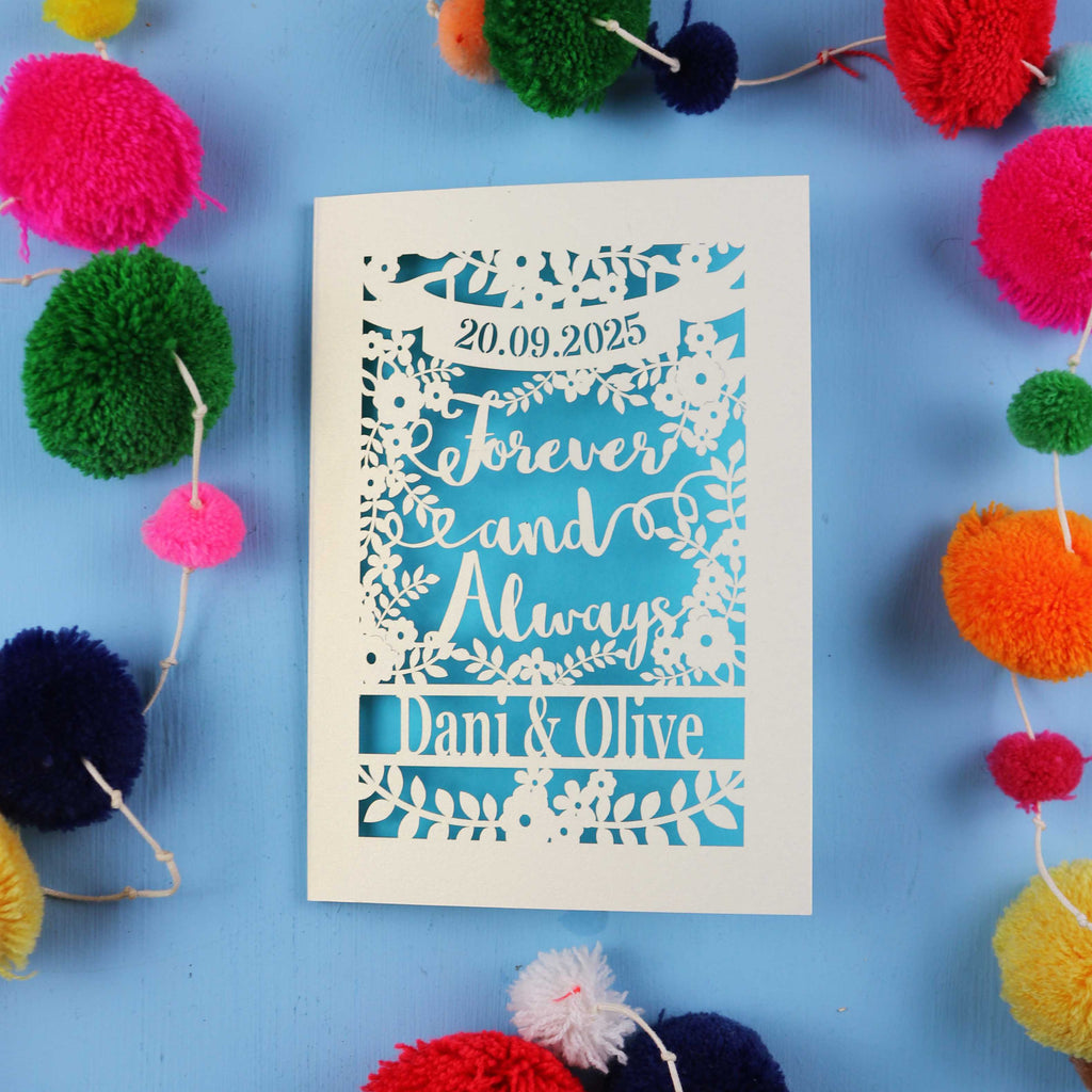 Decorative paper cut engagement card with personalized text on a blue background with colourful pom-poms.