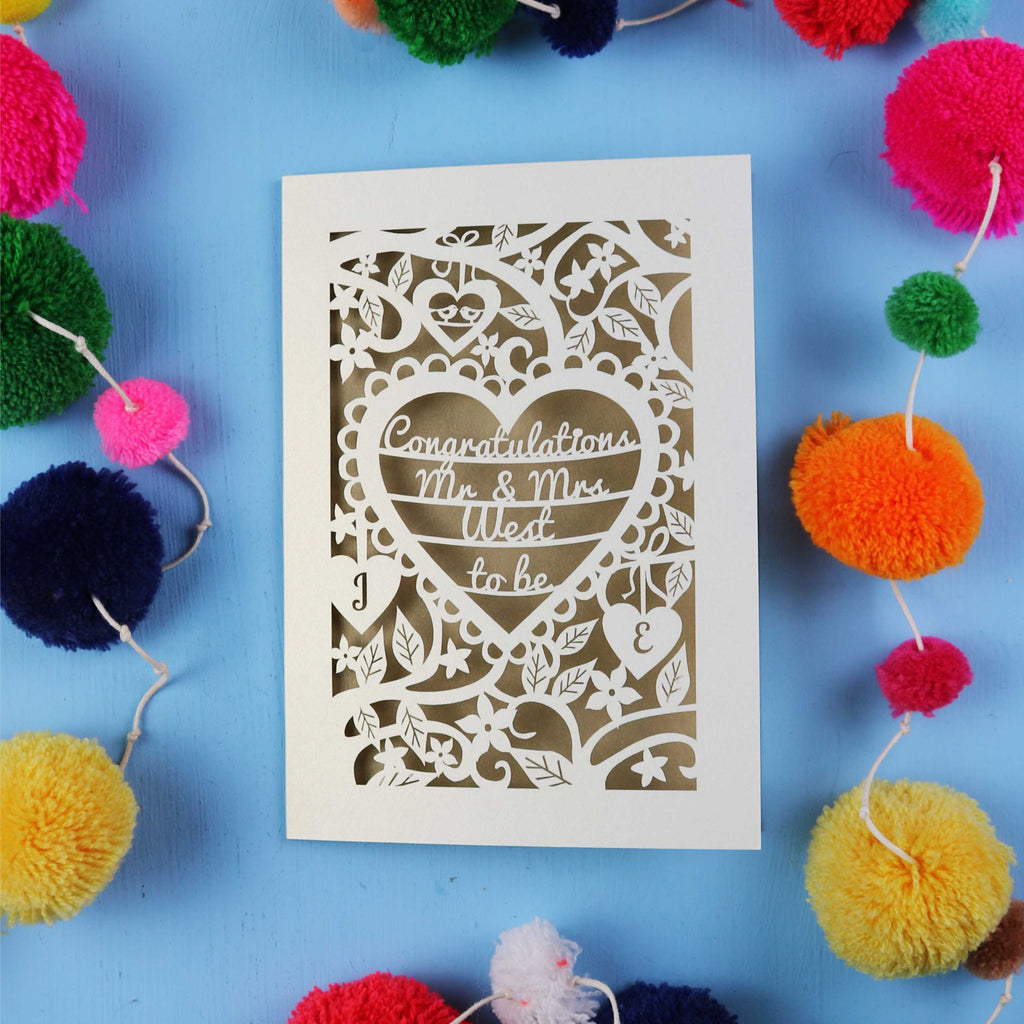 Laser-cut engagement card with decorative text on a blue background with colourful pom-poms.