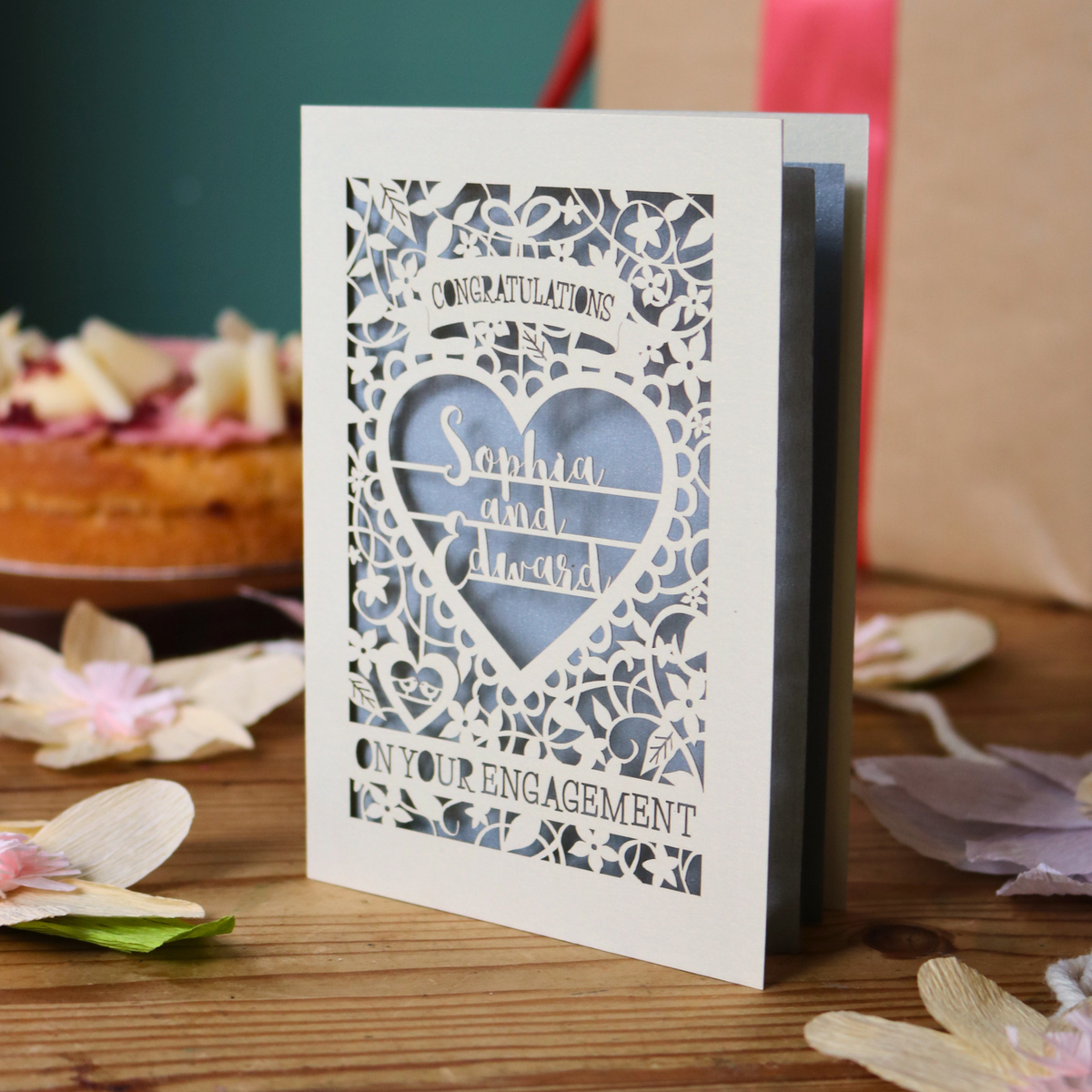 A cream laser cut engagement card with intricate floral border and heart design. Inside the heart are the names 'Sophia and Edward' in a brush script font, above is the word 'Congratulations', and below 'on your engagement'