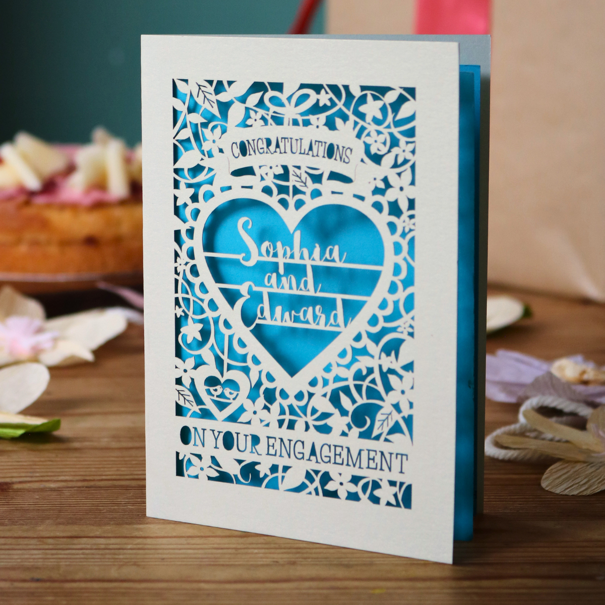 A cream laser cut engagement card with intricate floral border and heart design. Inside the heart are the names 'Sophia and Edward' in a brush script font, above is the word 'Congratulations', and below 'on your engagement'