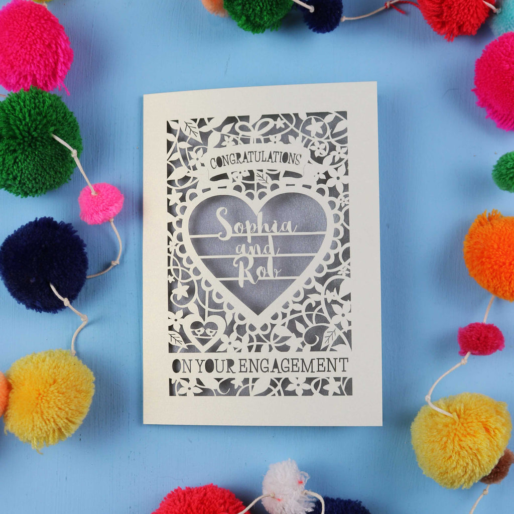 Engagement card with decorative heart design on a blue background with colourful pom-poms.