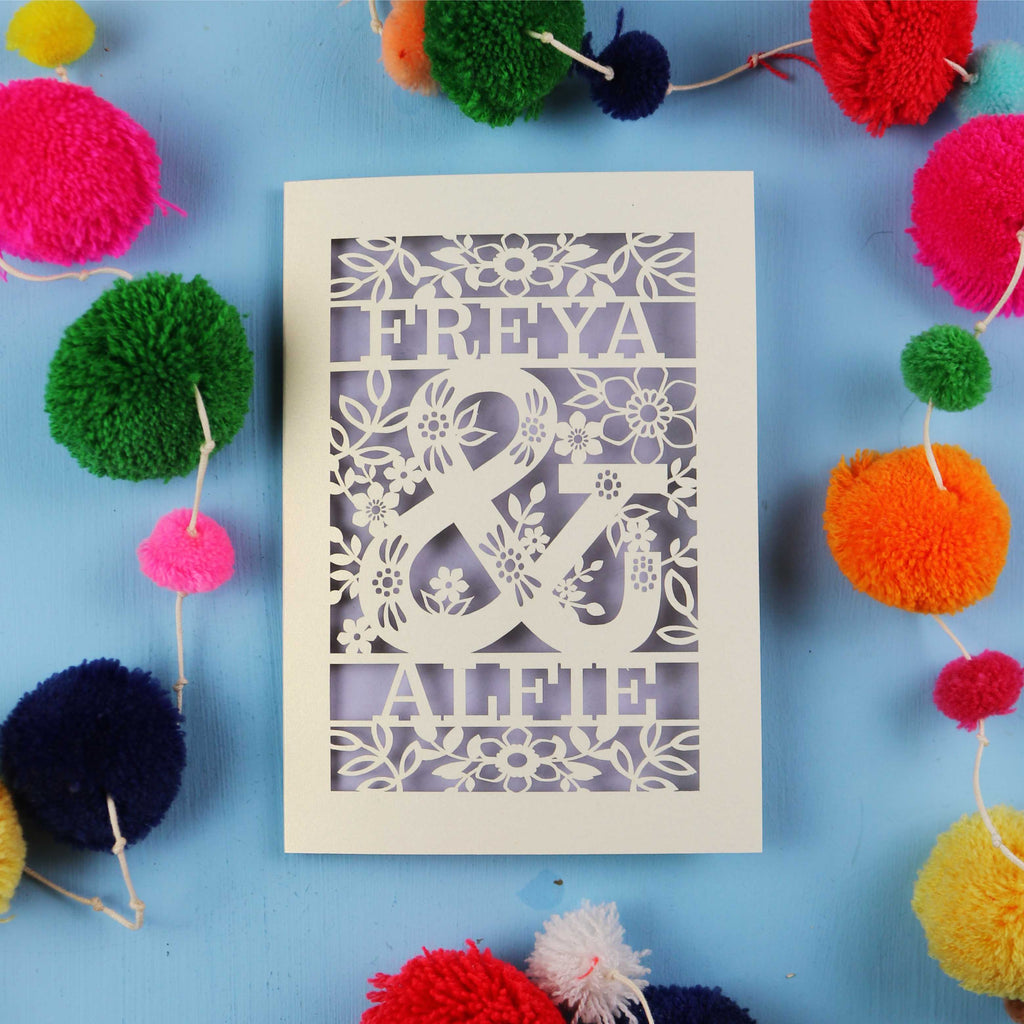 Decorative paper cut out engagement card with names 'FREYA' and 'ALFIE' surrounded by colourful pom-poms on a blue background