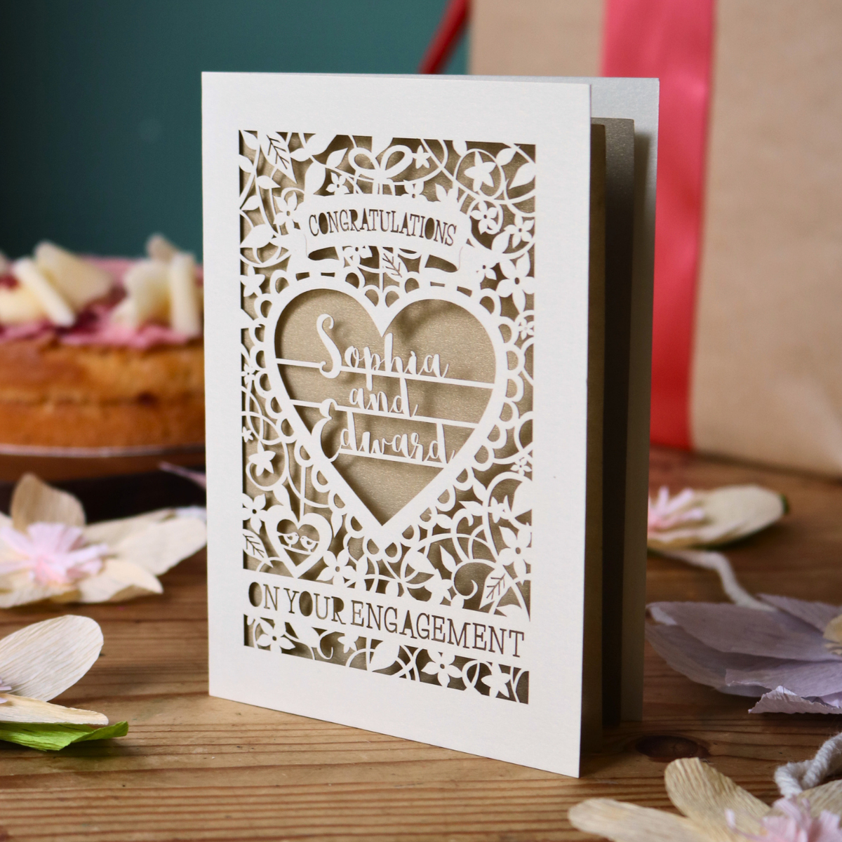 A cream laser cut engagement card with intricate floral border and heart design. Inside the heart are the names 'Sophia and Edward' in a brush script font, above is the word 'Congratulations', and below 'on your engagement'