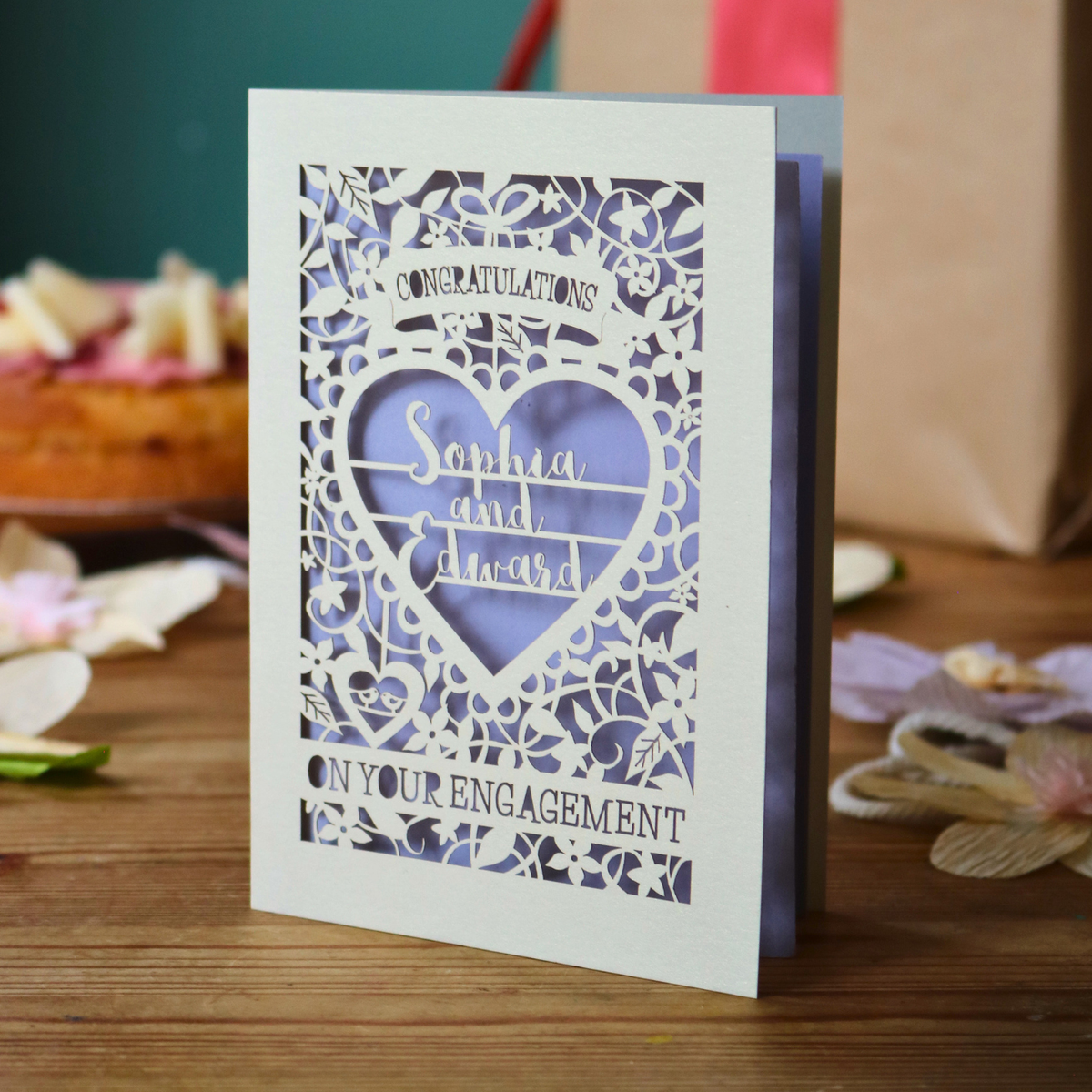 A cream laser cut engagement card with intricate floral border and heart design. Inside the heart are the names 'Sophia and Edward' in a brush script font, above is the word 'Congratulations', and below 'on your engagement'