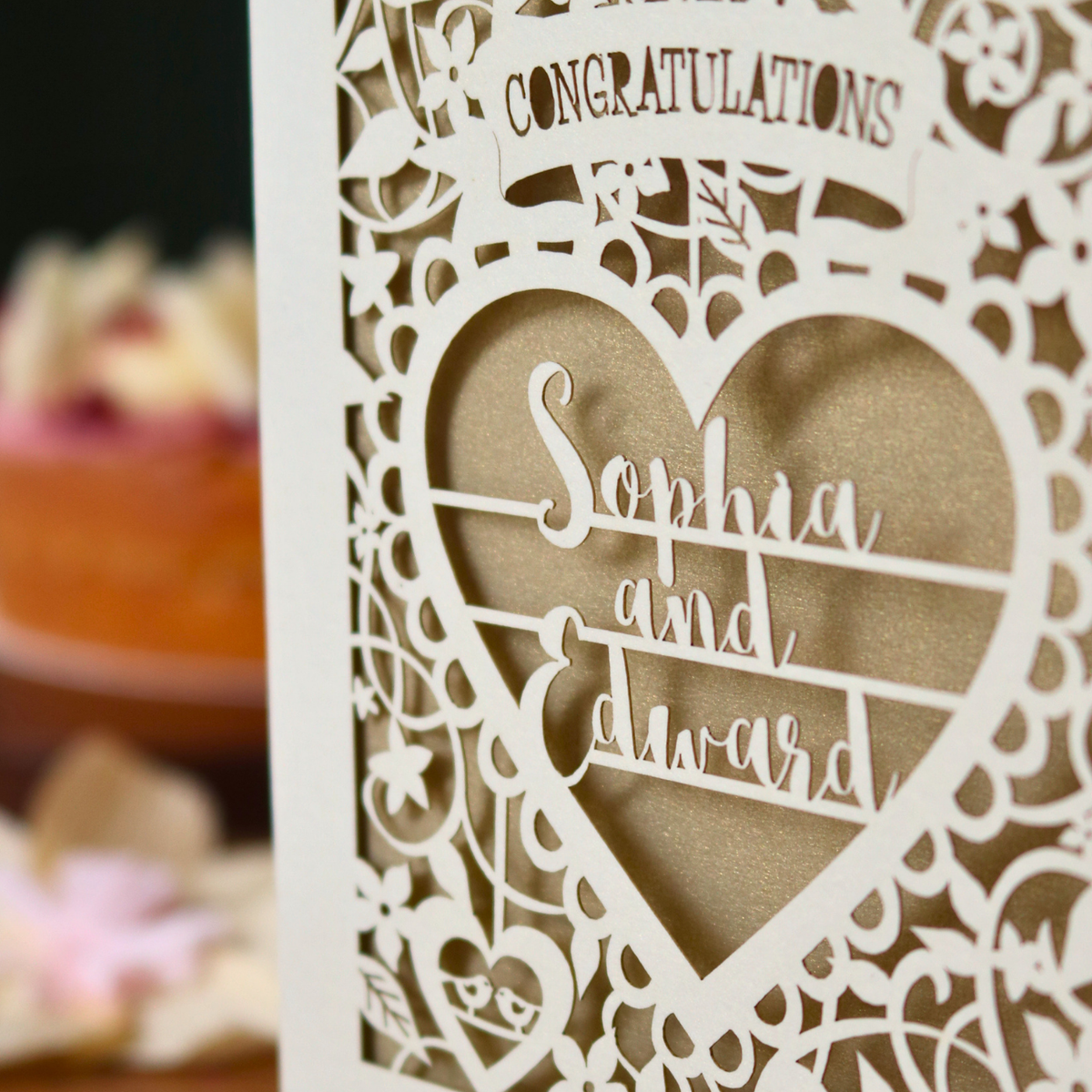 A close-up of a laser-cut congratulations card with intricate filigree patterns and a heart-shaped cut out containing the words 'Sophia and Edward' in elegant script over a gold glittery background.