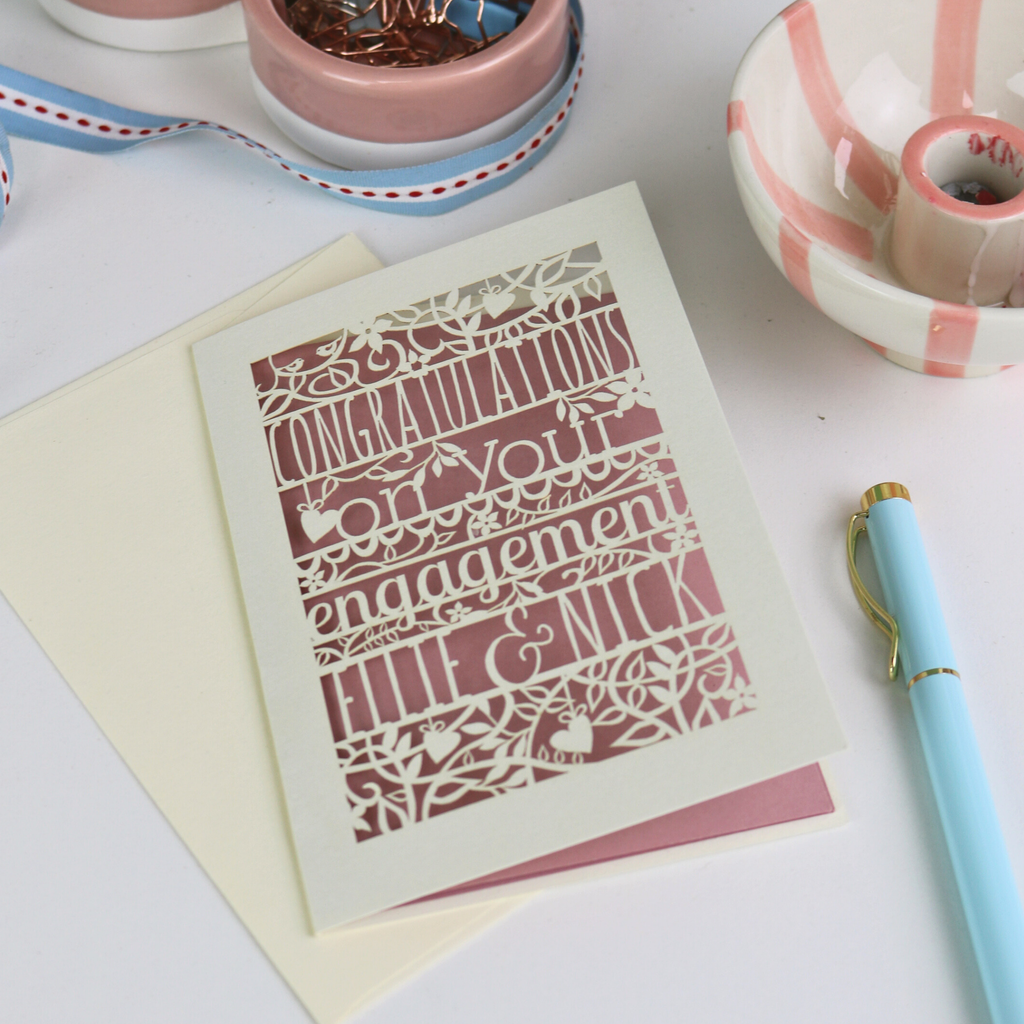 Engagement card with decorative text on a white surface with ribbons and a pen.
