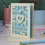 Keepsake laser cut new baby greeting card with cut-out designs on a desk with stationery items.