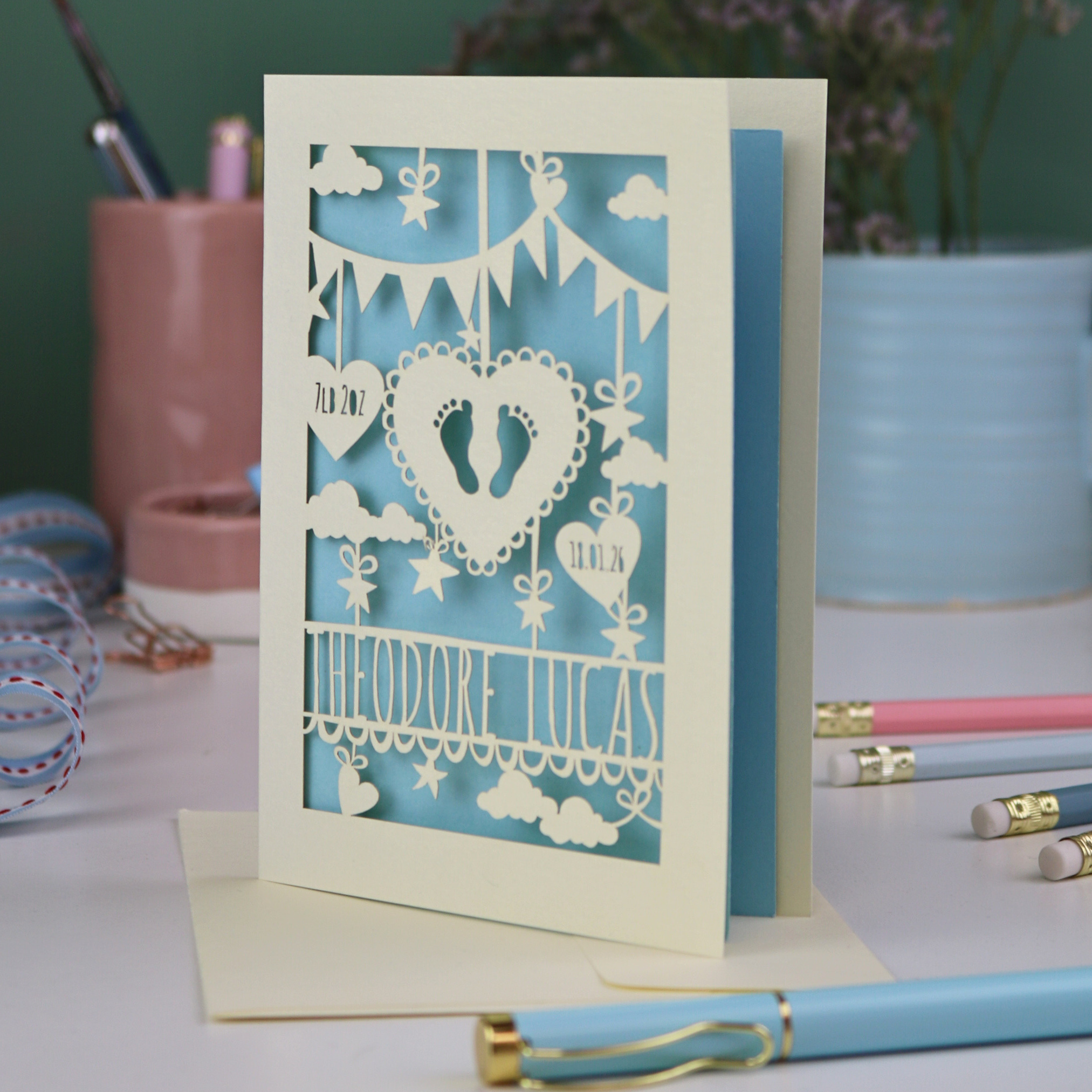 Keepsake laser cut new baby greeting card with cut-out designs on a desk with stationery items.