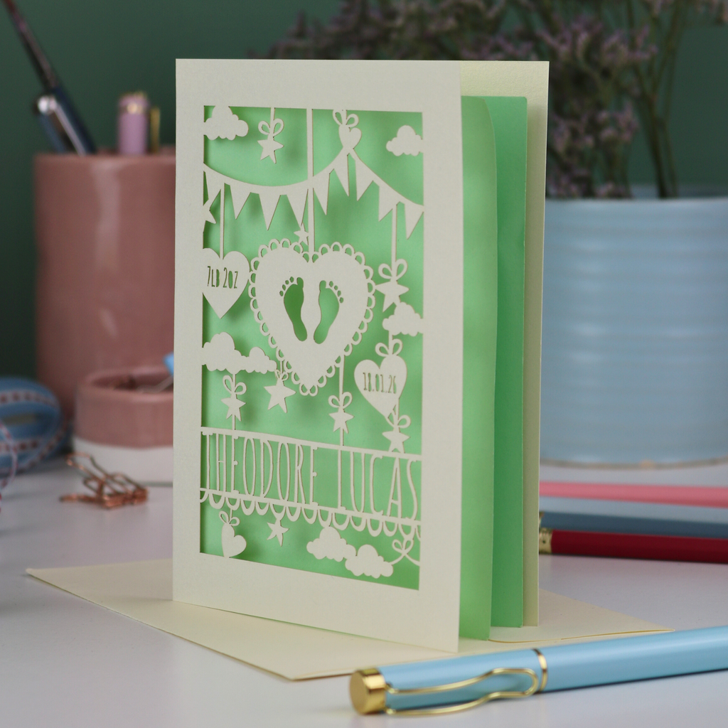 Decorative laser cut new baby greeting card with cut-out designs on a desk with stationery items.