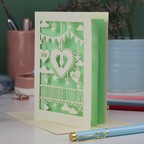 Decorative laser cut new baby greeting card with cut-out designs on a desk with stationery items.