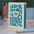 Personalised laser cut new baby greeting card with cut-out designs on a desk with stationery items.