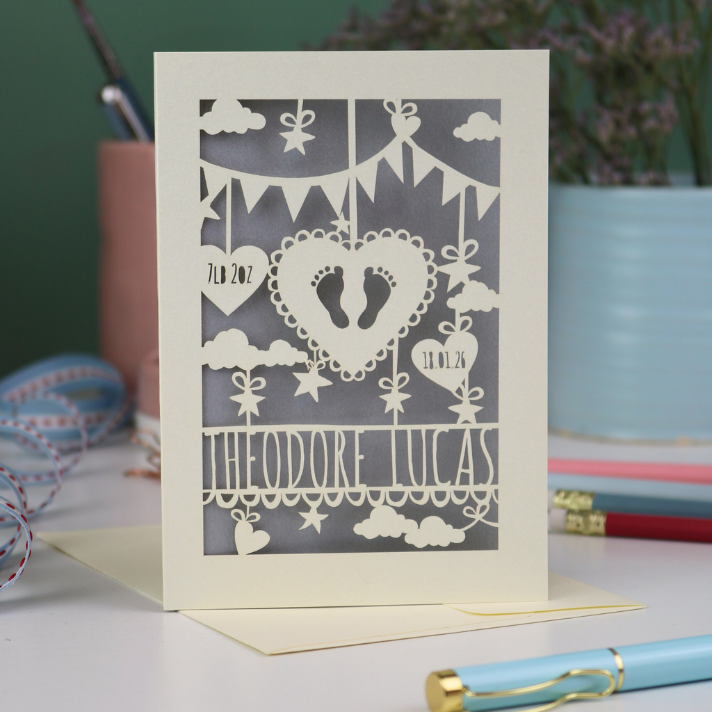 Decorative laser cut new baby greeting card with cut-out designs on a desk with stationery items.