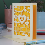 Decorative laser cut new baby greeting card with cut-out designs on a desk with stationery items.