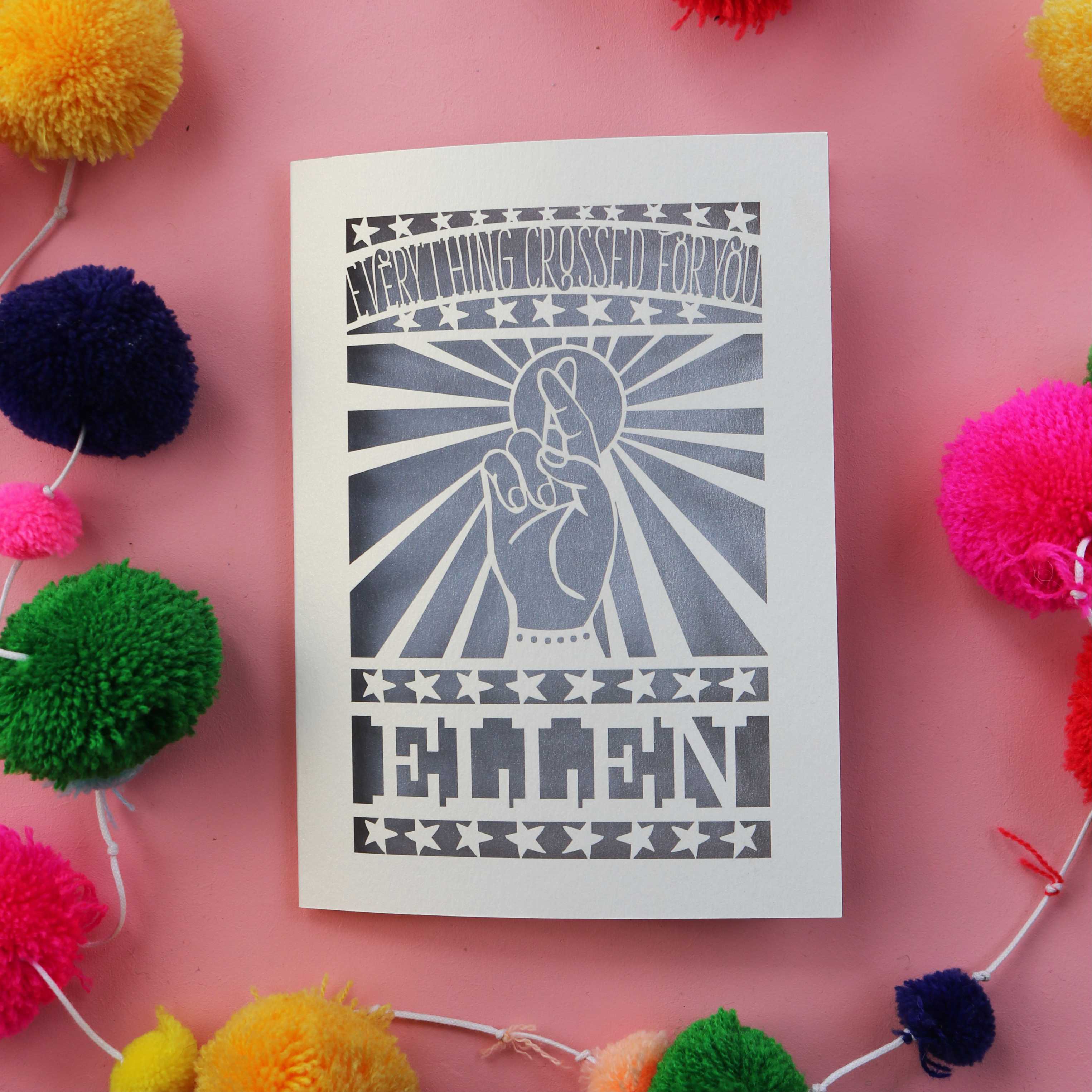 A paper cut good luck card personalised with a name and featuring a cut out hand with crossed fingers, surrounded by sunbeams