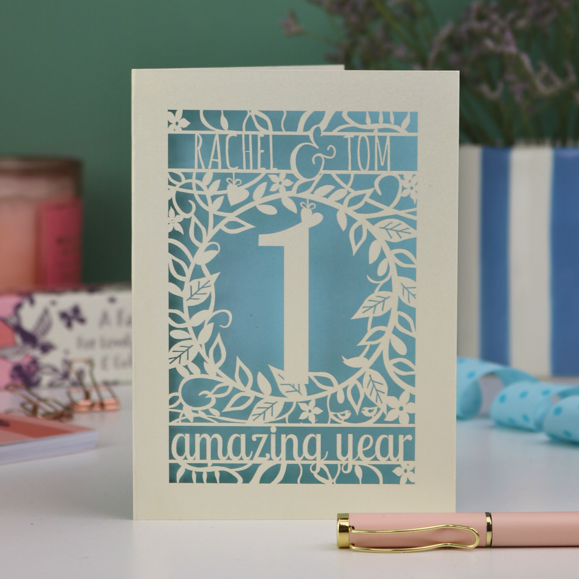 Laser cut first anniversary Card with '1 amazing year' design on a table with a pen and decorative items.
