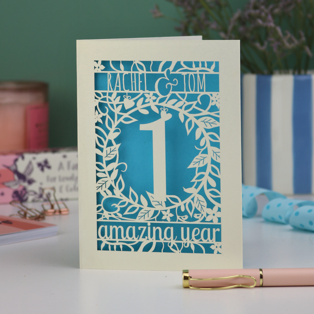Paper cut first anniversary Card with '1 amazing year' design on a table with a pen and decorative items.