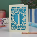 Paper cut first anniversary Card with '1 amazing year' design on a table with a pen and decorative items.