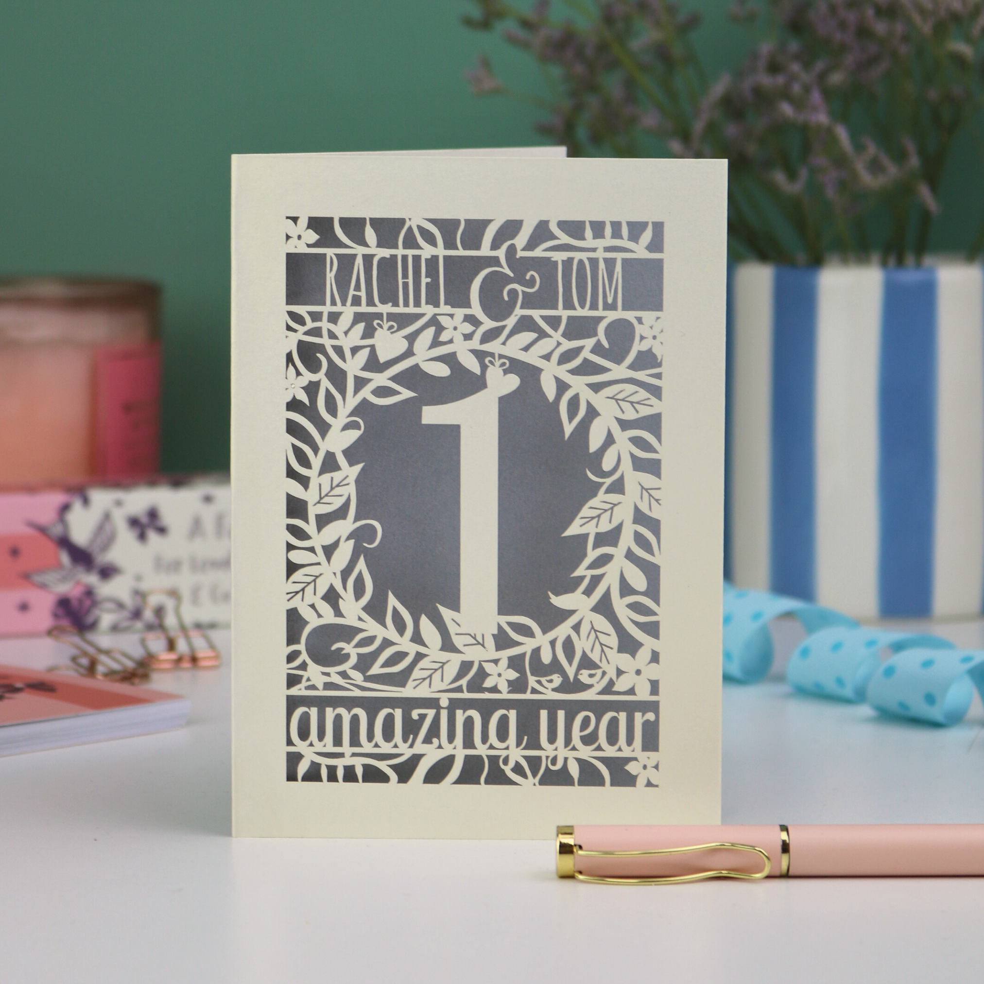 first anniversary Card with '1 amazing year' design on a table with a pen and decorative items.