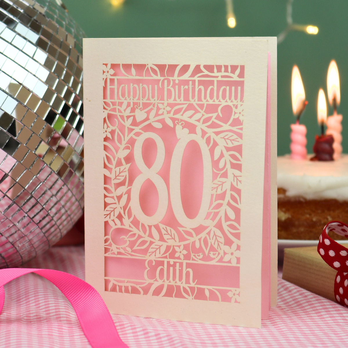 A pink and cream laser-cut 80th birthday card standing on a pink-and-white checked tablecloth, surrounded by party props including a disco ball, lit candles, and a cake in the background.
