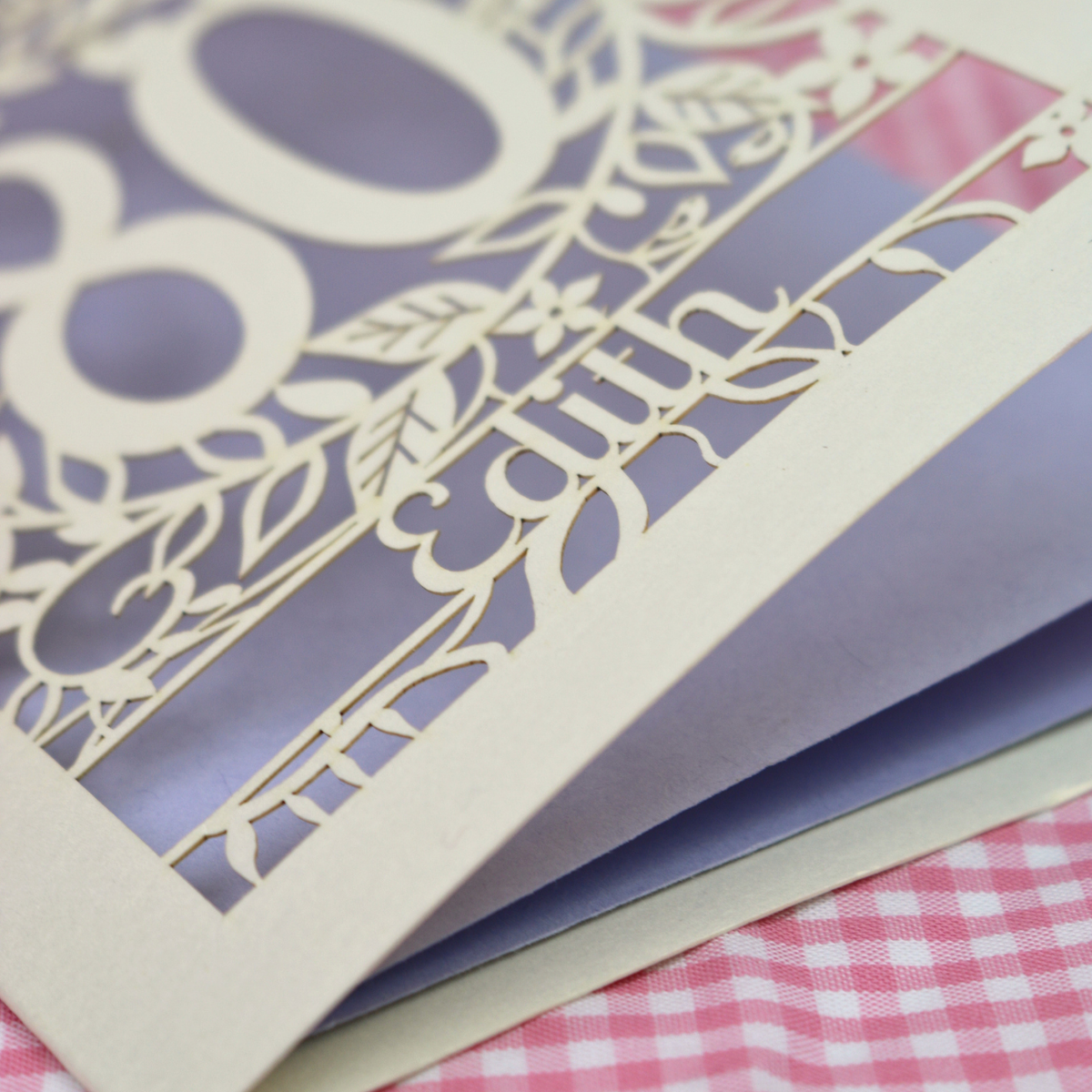 A close-up of a laser-cut birthday card with ornate floral and script cut outs, layered over a purple backing, resting on pink and white checked fabric.