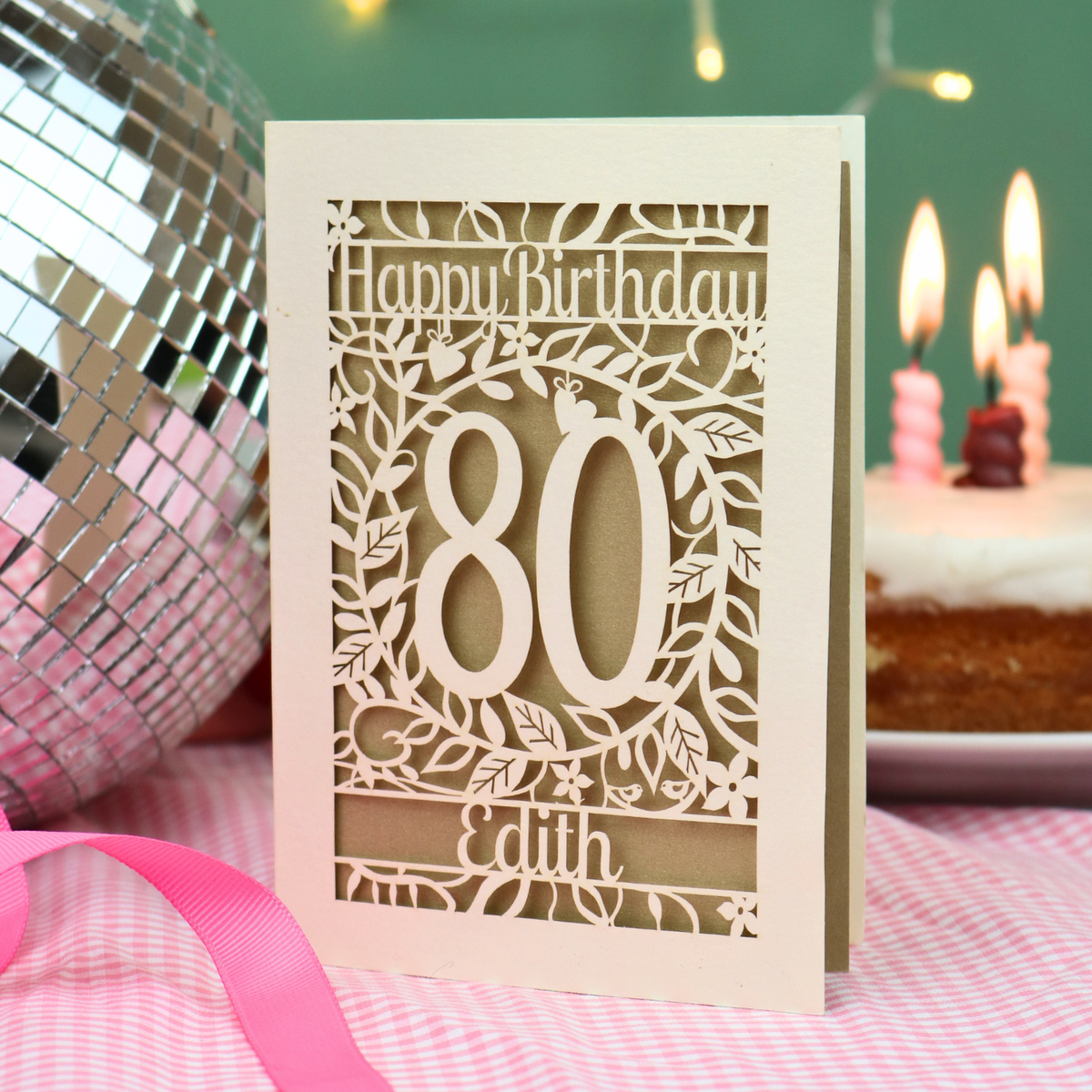 A personalized laser-cut birthday card with 'Happy Birthday', the number '80', and the name 'Edith', displayed on a pink checked tablecloth next to a pink ribbon, a disco ball, and a birthday cake in the background.