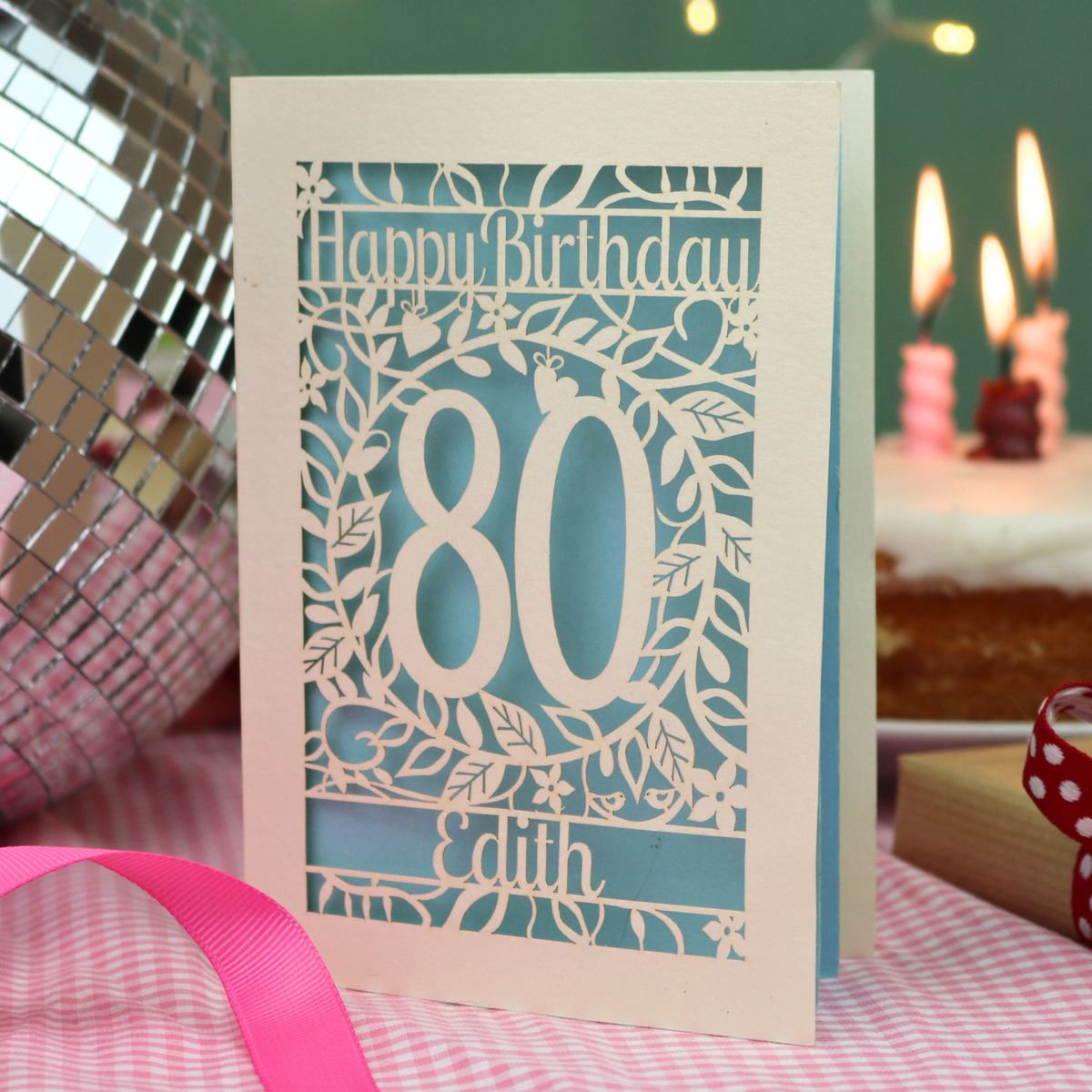 A laser-cut 80th birthday card with a cream-colored border and light blue inner paper, featuring the text 'Happy Birthday' and the name 'Edith' surrounded by floral designs. The card is displayed on a pink gingham tablecloth with a pink ribbon, a mirror, and a blurred birthday cake in the background.