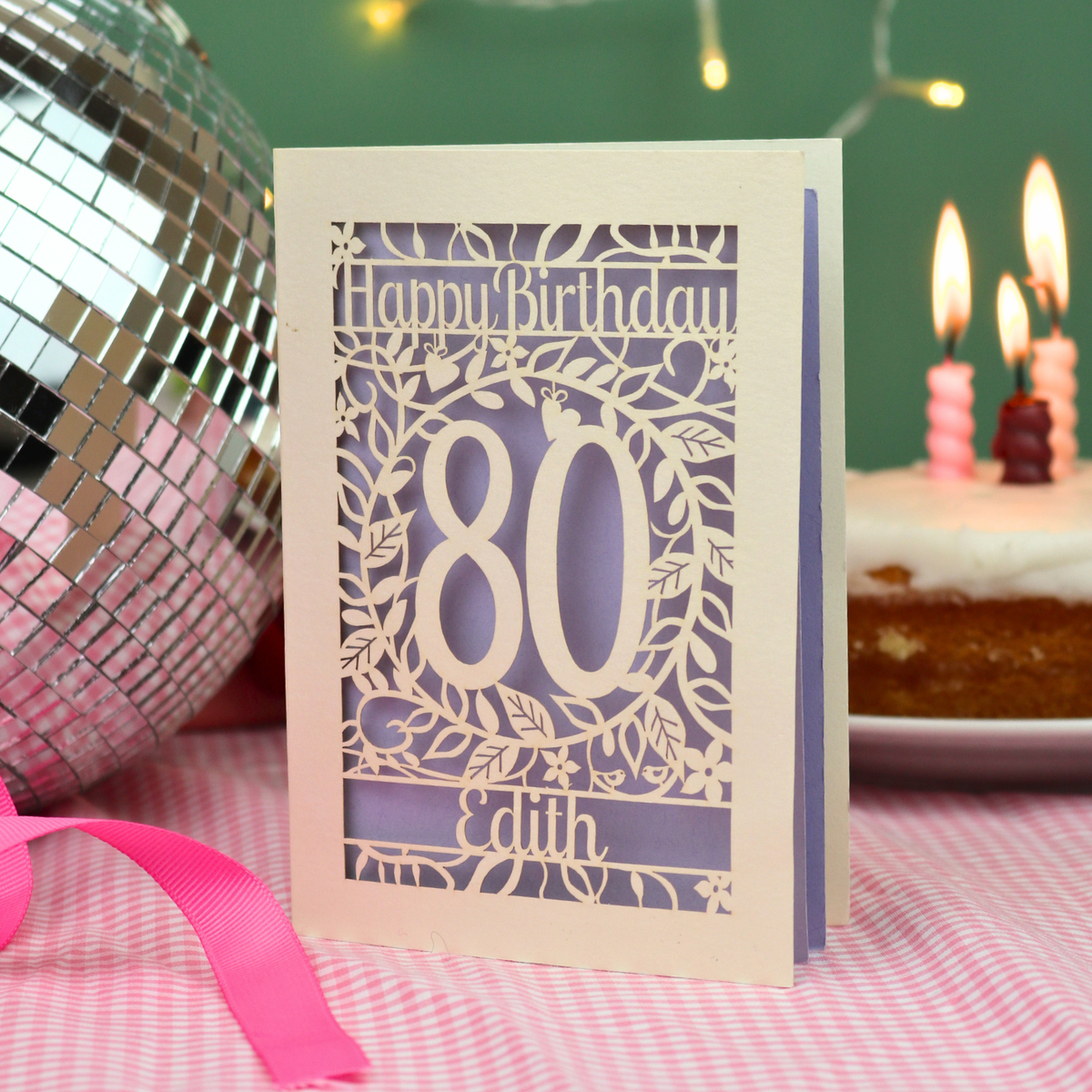 A personalised laser-cut birthday card with 'Happy Birthday', the number '80', and the name 'Edith', displayed on a pink checked tablecloth next to a pink ribbon, a disco ball, and a birthday cake in the background.