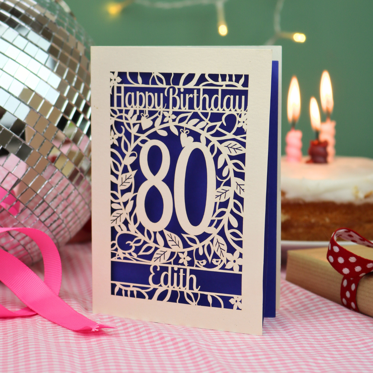 A personalized laser-cut birthday card with 'Happy Birthday', the number '80', and the name 'Edith', displayed on a pink checked tablecloth next to a pink ribbon, a disco ball, and a birthday cake in the background.