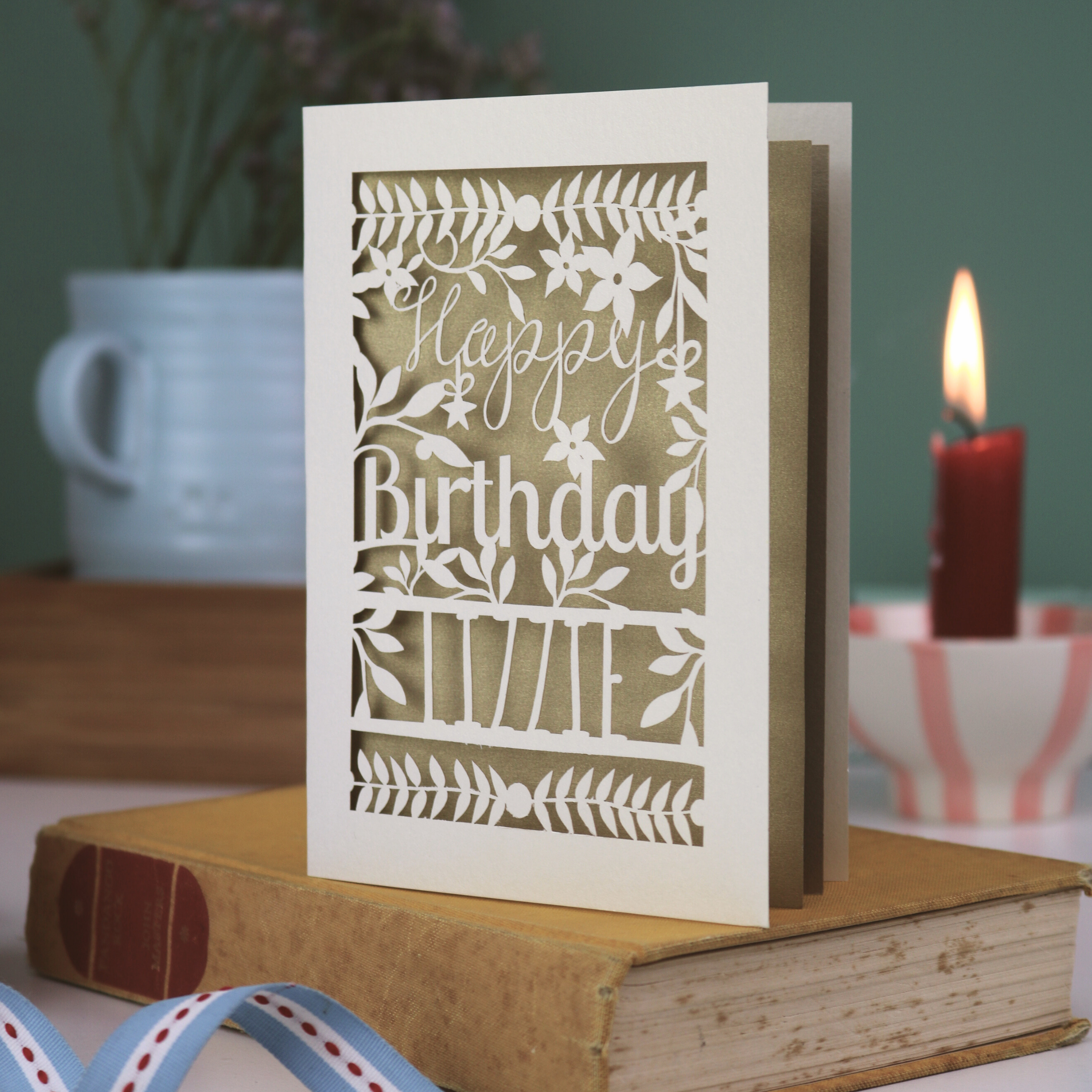 Birthday card with floral design and 'Happy Birthday' text on a book with a candle and plant in the background.