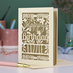 A cream laser cut birthday card with text "Happy Birthday" and personalised with the name 'Lizzie', on a desk with pen and stationery items