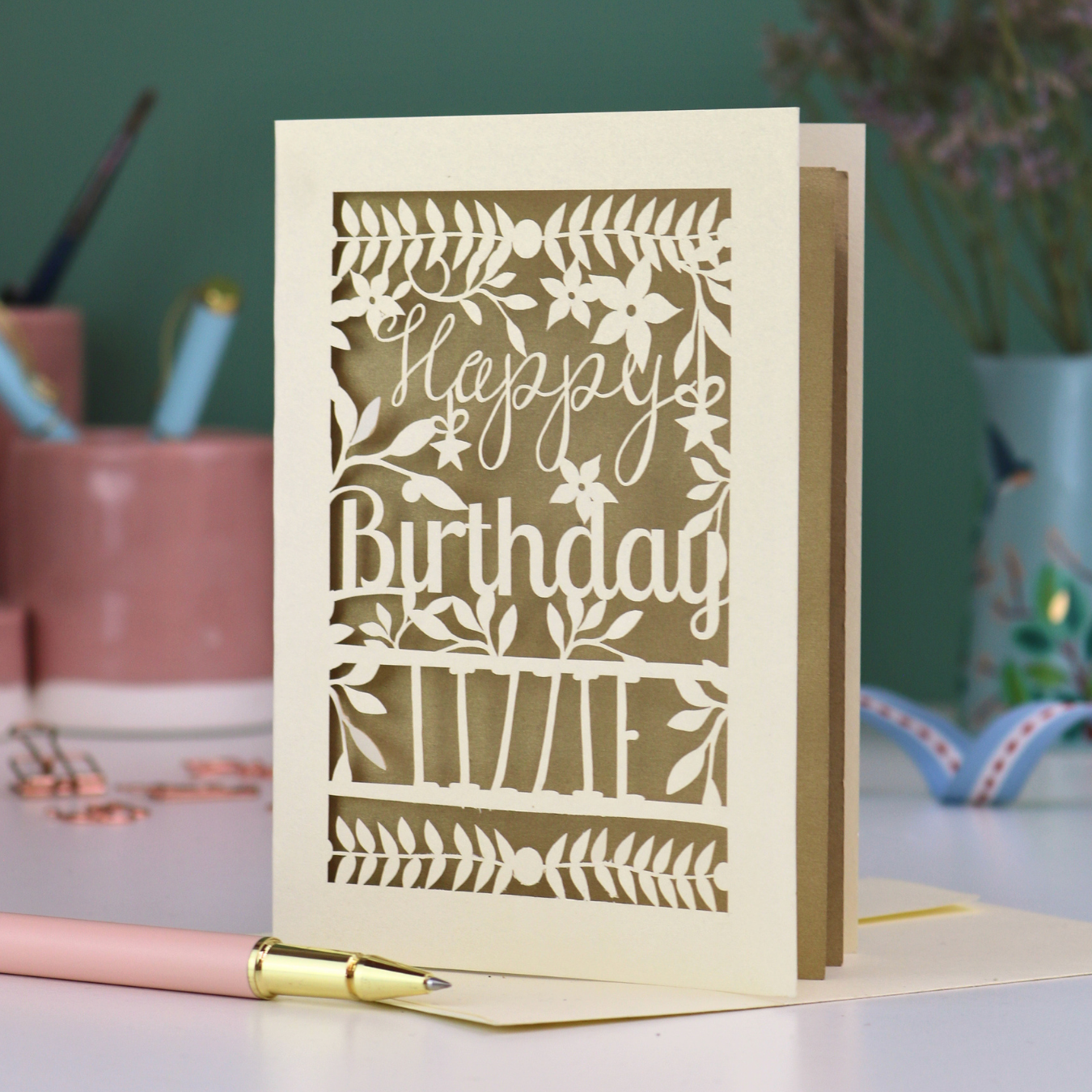 A cream laser cut birthday card with text "Happy Birthday" and personalised with the name 'Lizzie', on a desk with pen and stationery items