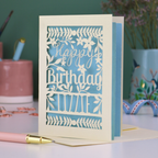 A cream laser cut birthday card with text "Happy Birthday" and personalised with the name 'Lizzie', on a desk with pen and stationery items