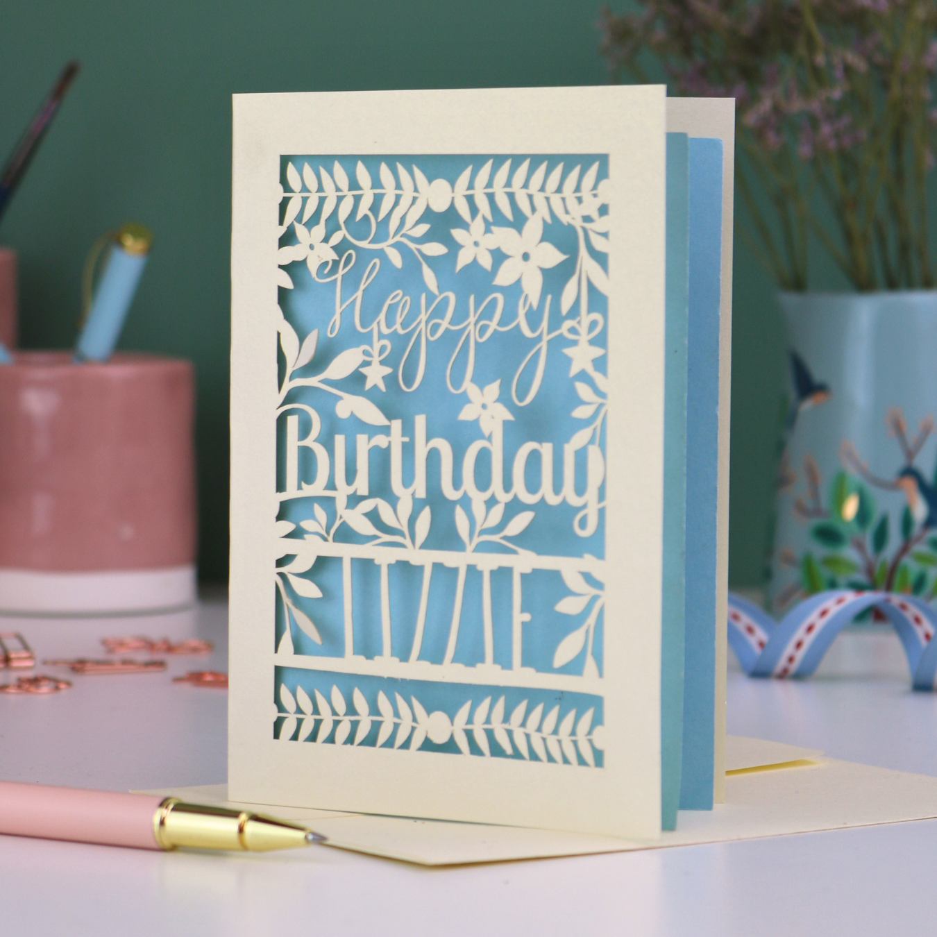 A cream laser cut birthday card with text "Happy Birthday" and personalised with the name 'Lizzie', on a desk with pen and stationery items