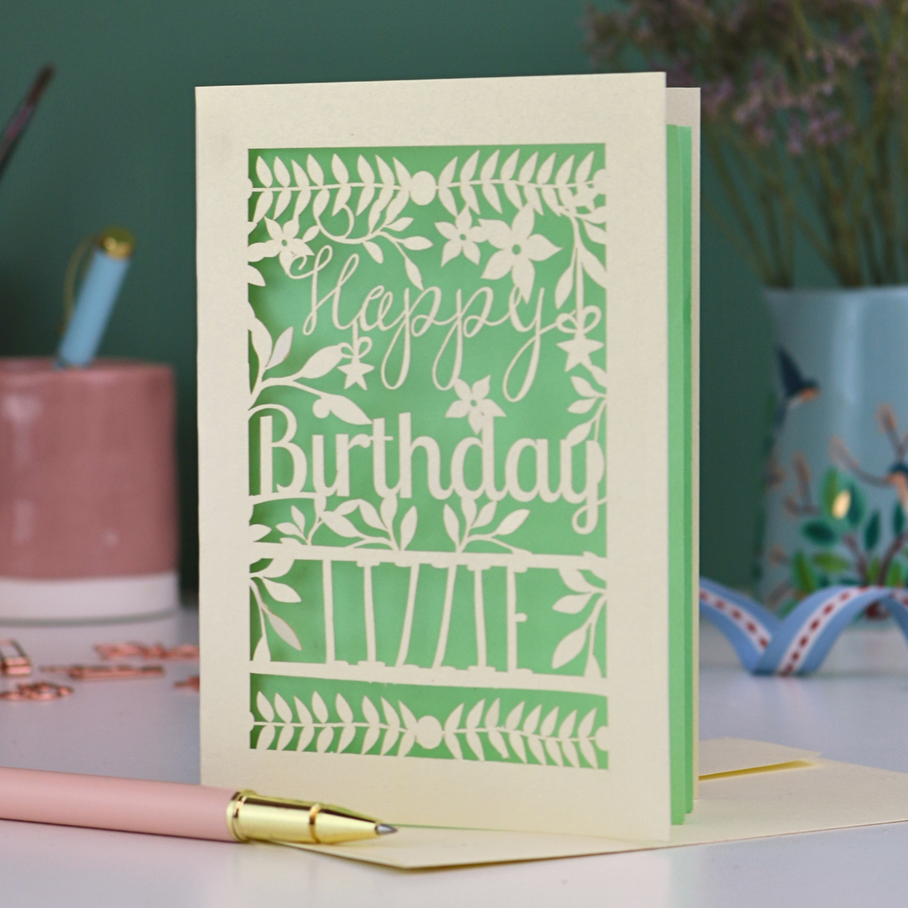 A cream laser cut birthday card with text "Happy Birthday" and personalised with the name 'Lizzie', on a desk with pen and stationery items