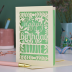 A cream laser cut birthday card with text "Happy Birthday" and personalised with the name 'Lizzie', on a desk with pen and stationery items