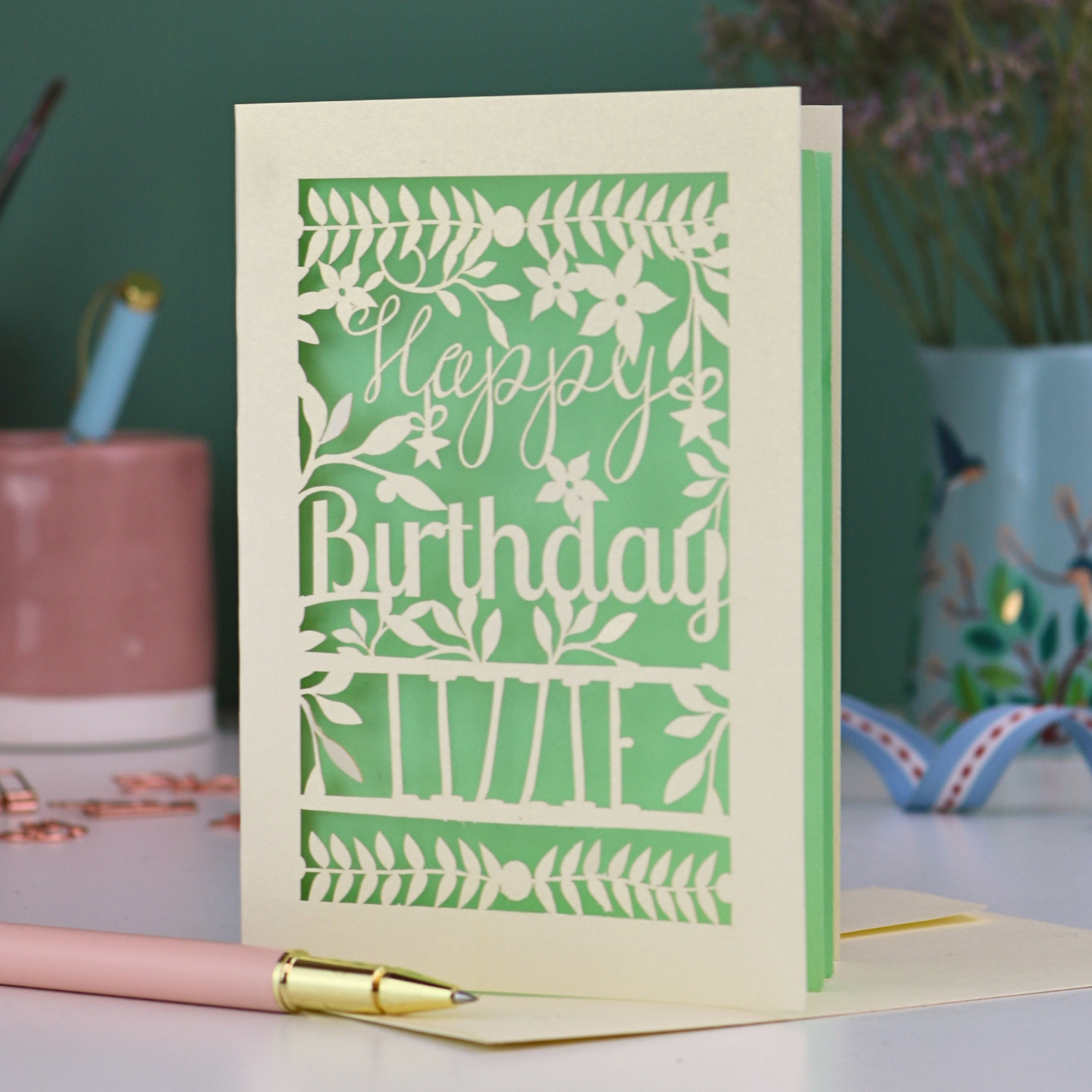 A cream laser cut birthday card with text "Happy Birthday" and personalised with the name 'Lizzie', on a desk with pen and stationery items