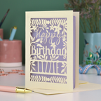 A cream laser cut birthday card with text "Happy Birthday" and personalised with the name 'Lizzie', on a desk with pen and stationery items