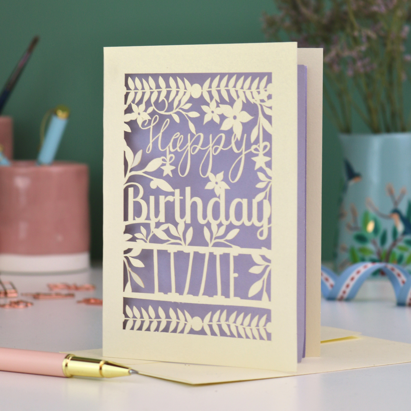 A cream laser cut birthday card with text "Happy Birthday" and personalised with the name 'Lizzie', on a desk with pen and stationery items