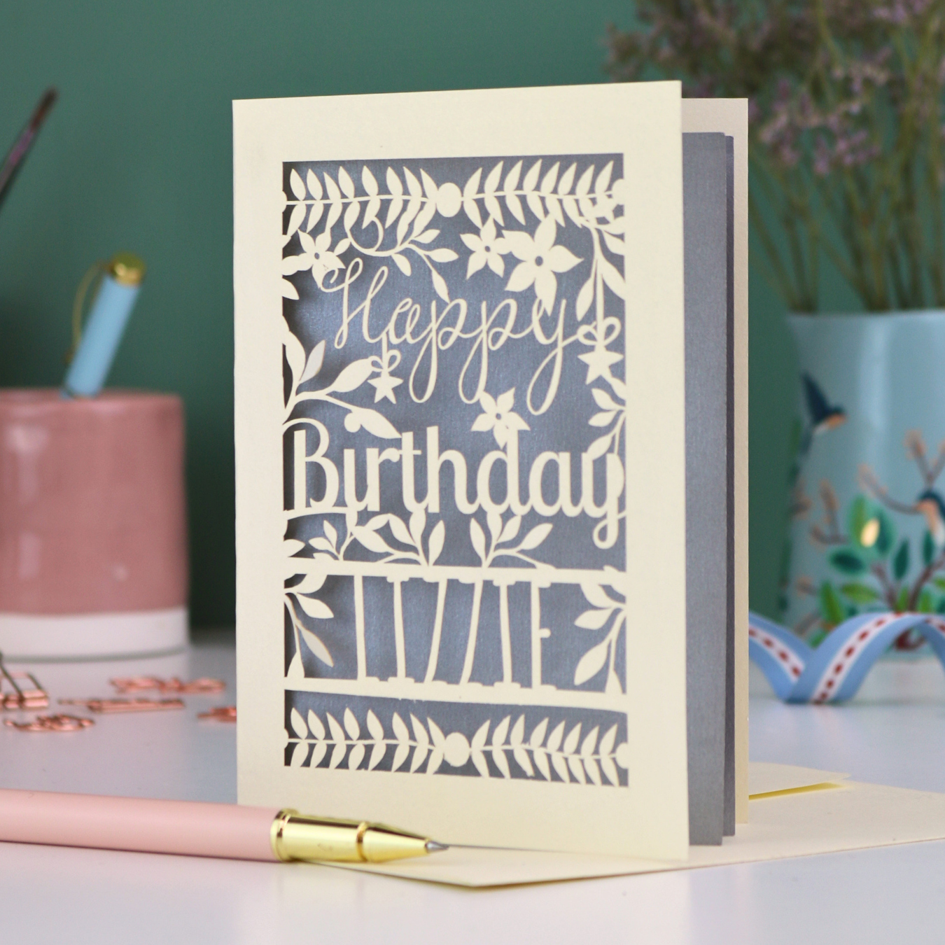 A cream laser cut birthday card with text "Happy Birthday" and personalised with the name 'Lizzie', on a desk with pen and stationery items