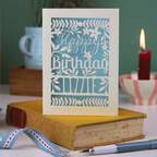 Birthday card with floral design and 'Happy Birthday' text on a book with a candle and plant in the background.