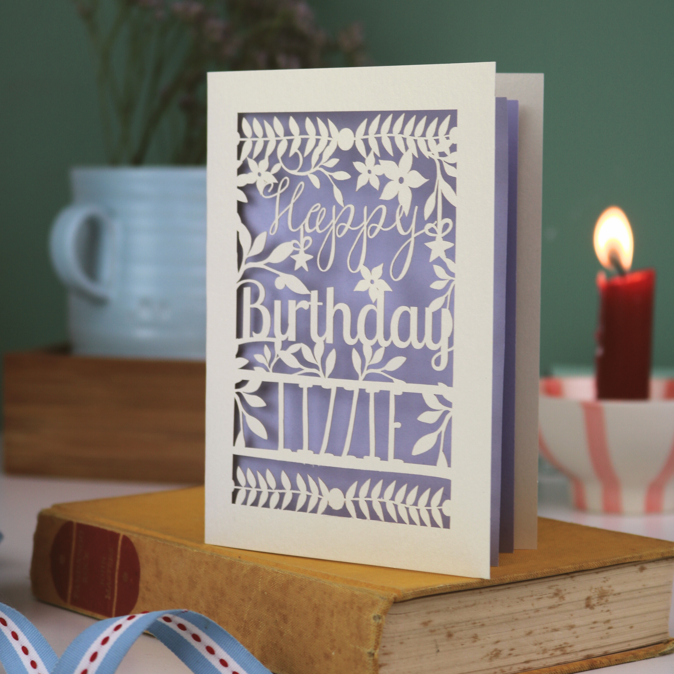 Birthday card with floral design and 'Happy Birthday' text on a book with a candle and plant in the background.
