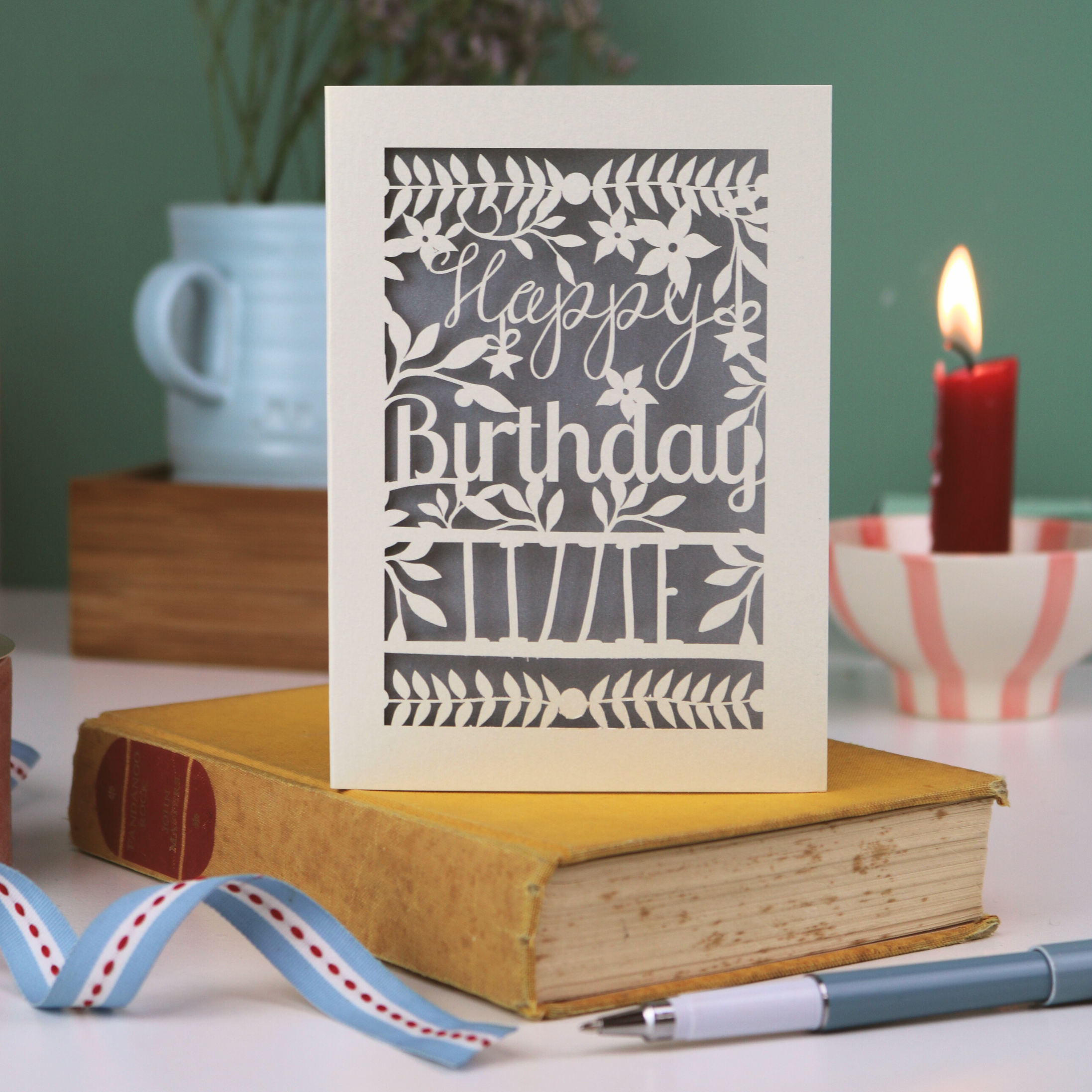 Birthday card with floral design and 'Happy Birthday' text on a book with a candle and plant in the background.