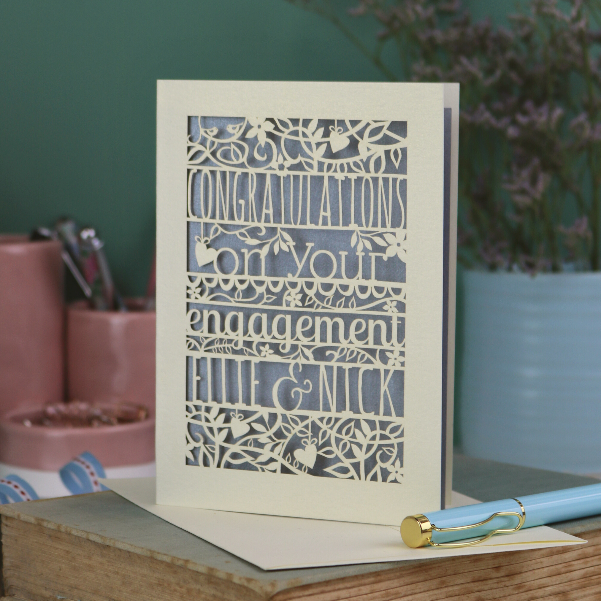 Engagement card with decorative text on a wooden surface with stationery items.